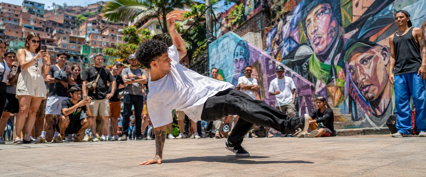 Young breakdancer performing in Comuna 13 plaza surrounded by murals and a crowd of tourists and locals